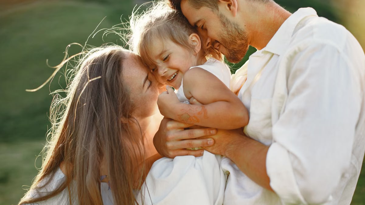 A joyful family moment with parents embracing their child outdoors in warm sunlight.
